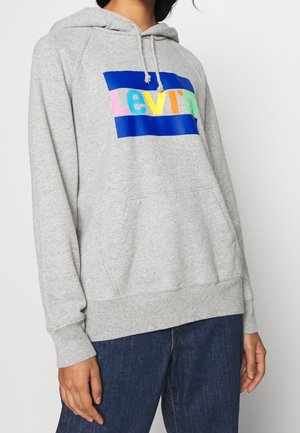 Personne portant un sweat à capuche gris clair avec un logo Levi's multicolore, capuche à cordon, poche kangourou et jean bleu foncé.