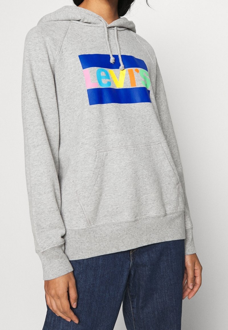 Personne portant un sweat à capuche gris clair avec un logo Levi's multicolore, capuche à cordon, poche kangourou et jean bleu foncé.