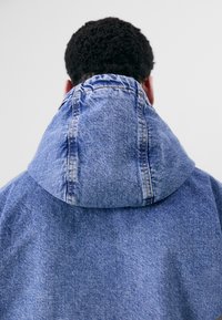 Džínová bunda s velkou složenou kapucí; má viditelné stehy a strukturovanou modrou látku. Pohled zezadu.