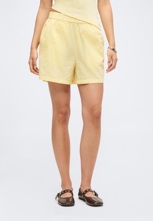 Vrouw draagt gele geruite shorts, een lichtgeel top, een zilveren armband en donkerbruine Mary Jane schoenen, staand op een witte achtergrond.