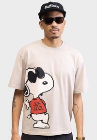 Heledad beež puuvillane T-särk, millel on suur graafika Snoopy'st, kes kannab prille ja punast "JOE COOL" särk. Lühikesed varrukad ja lõdva lõikega.