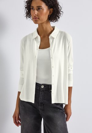 Cárdigan blanco de botones hecho de tela suave, con cuello, mangas largas y un ajuste relajado, combinado con una camiseta sin mangas blanca y jeans oscuros.