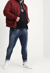 Rode bomberjack over een zwarte capuchontrui met afbeelding, versleten blauwe jeans en witte sneakers. Details van een casual outfit.