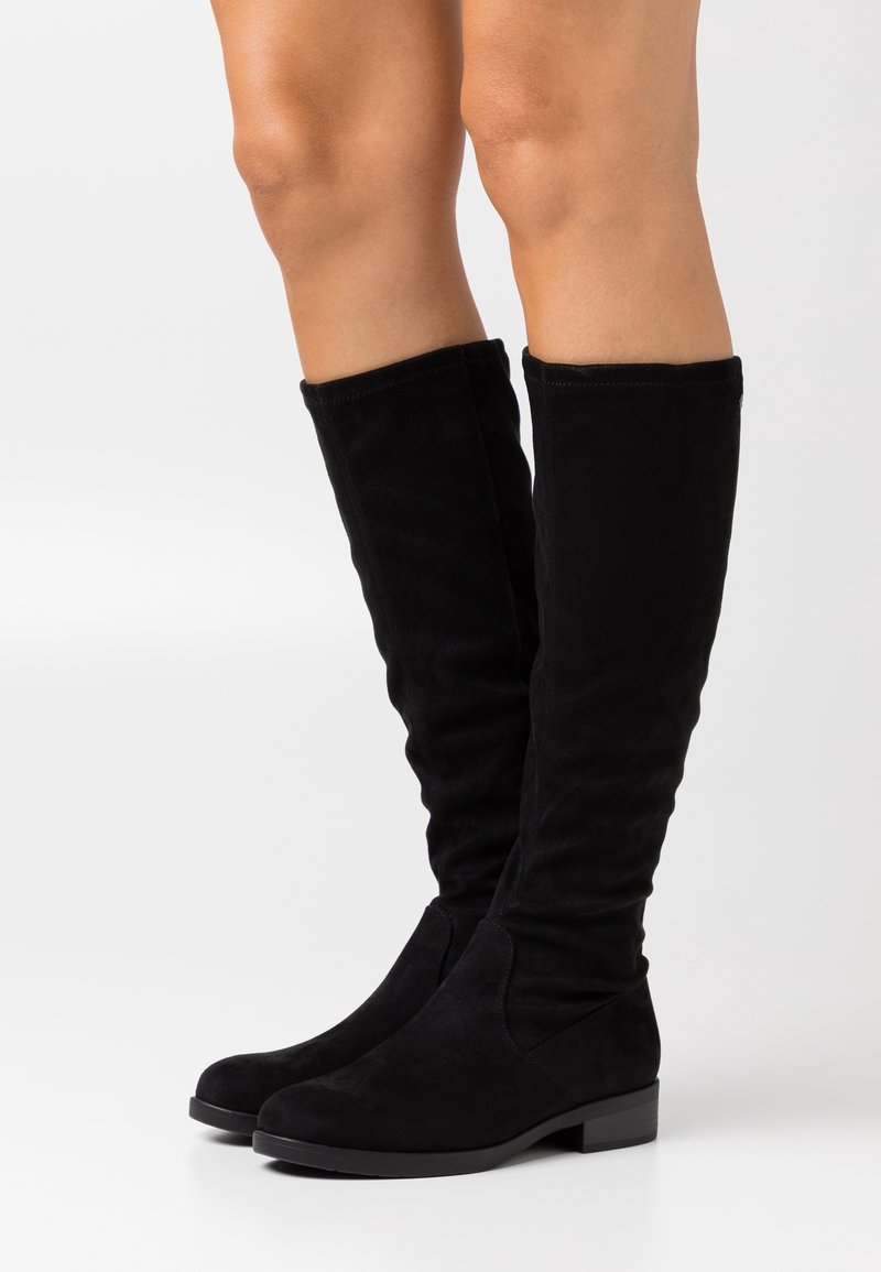 Bottes noires en daim montant jusqu'aux genoux portées sur des jambes nues, semelle plate avec petit talon, design simple, finition lisse sur fond blanc.