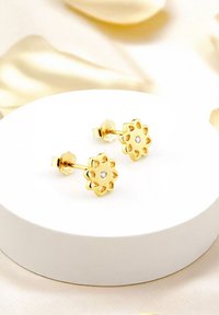 Goldene blumenförmige Ohrstecker mit ausgeschnittenen Blütenblättern und einem kleinen zentralen Stein. Schlankes Design auf einer weißen runden Präsentationsbasis.