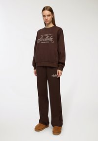 Brązowa bluza z kapturem i pasujące spodnie z haftowanymi grafikami "Stockholm Leisure Club". Miękka tkanina, luźny krój, okrągły dekolt i szerokie nogawki.