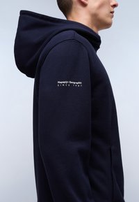 Felpa con cappuccio blu navy dalla texture morbida, con un logo sulla manica che recita "Napapijri Geographic Since 1987".