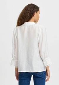 White, long-sleeve blouse with textured fabric, puffed sleeves, and lace trim at cuffs. Features a classic collar and loose fit.