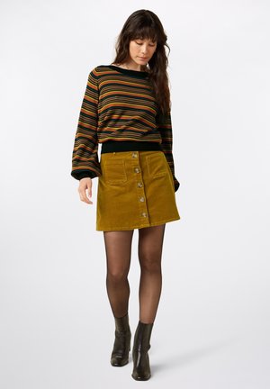 Gestreepte trui in meerdere kleuren met lange mouwen, gecombineerd met een mosterdkleurige corduroy rok met een knopenfront en twee zakken. Zwarte enkellaarsjes.