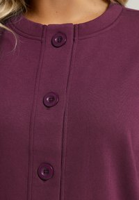 Lila bomullsplagg med rund halsringning, framknäppning med tre matchande knappar och en slät, strukturerad tygkvalitet.