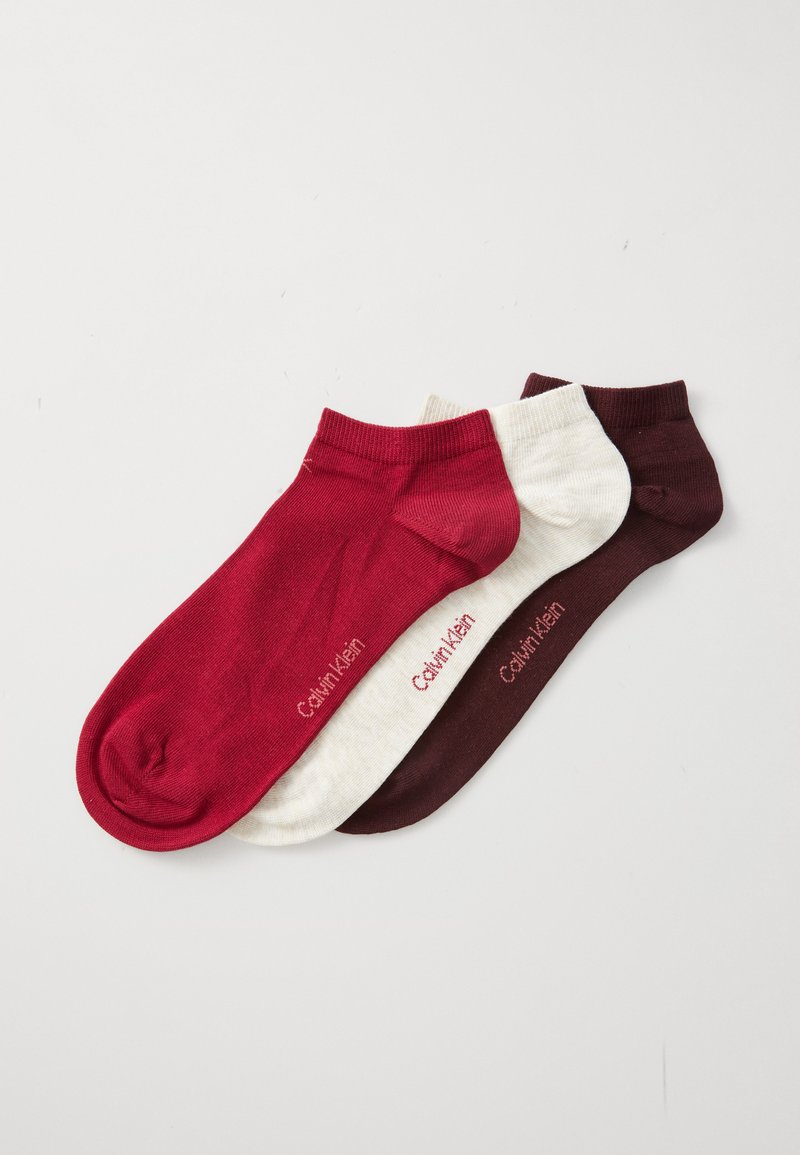 Tre låga strumpor i röd, krämfärgad och mörkbrun färg, var och en märkt med "Calvin Klein" på en vit bakgrund.
