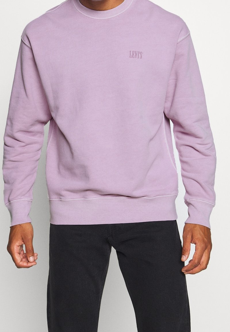 Homme portant un sweat-shirt Levi's violet clair avec un pantalon noir, debout devant un fond blanc.