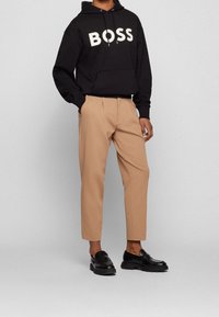 Persona che indossa pantaloni cropped beige con mocassini in pelle nera e felpa con cappuccio nera con la scritta "BOSS" sul petto, con le mani nelle tasche.
