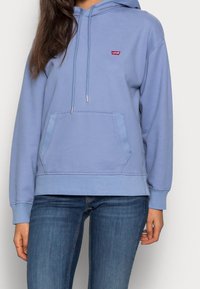 Kvinna i ljusblå Levi's hoodie med framficka och jeans, som står mot en enfärgad vit bakgrund.