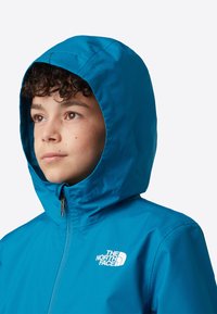 Jachetă impermeabilă de culoare verde-albăstrui cu fermoar frontal și glugă. Prezintă un logo alb pe piept și o textură netedă, strălucitoare.