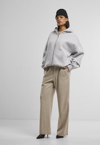 Graue Zip-Hoodie mit Fronttaschen, kombiniert mit beigen, weiten Hosen. Das Model trägt eine schwarze Mütze und spitze schwarze High Heels.