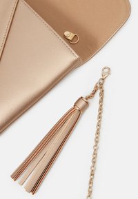 Metallisch goldfarbene Clutch-Tasche mit glatter Oberfläche, Druckverschluss und angehängtem Quast mit Ketten-Detail. Elegantes und kompaktes Design.
