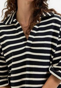 Femme portant une chemise à rayures horizontales noir et blanc avec des manches amples et des cheveux bruns bouclés partiellement visibles.