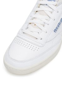 Tenis cu piele albă, având o textură netedă, cutie de degete perforată, accent de logo albastru și șireturi plate; talpa rotundă din cauciuc este vizibilă.