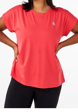 Kobieta w luźnej czerwonej koszulce z krótkim rękawem i czarnych legginsach, z rękami opartymi na biodrach, na białym tle.