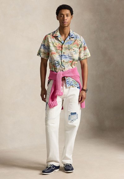 Mladý muž, ktorý má na sebe kvetovanú krátkoročnú košeľu, ružový sveter zviazaný v páse, biele rifle s náplasťami a námornícke modré lodné topánky na neutrálnom pozadí.