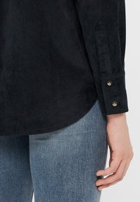 Svart kordfløyels skjorte med lange ermer og rundet bunnkant, med knapper på ermene, i en myk tekstur, kombinert med lyseblå denimjeans.