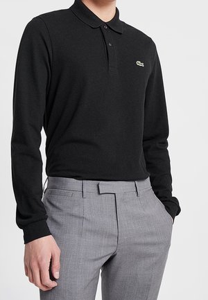 Polo noir à manches longues avec col à trois boutons et logo vert. Associé à un pantalon tailleur gris clair avec poches avant.