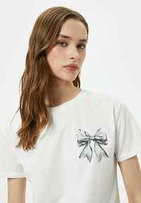 Hvid bomulds-T-shirt med et trykt sort og sølv bånddesign på venstre brystområde. Korte ærmer og rund halsudskæring.