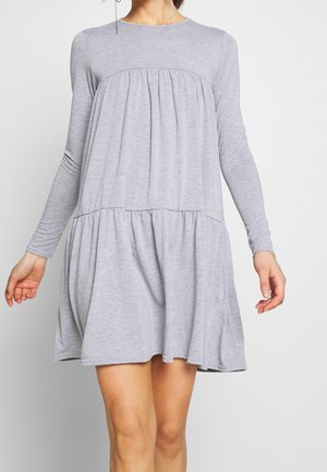 Femme portant une robe grise claire à manches longues, ample, au-dessus du genou, avec des coutures froncées au niveau de la poitrine et des hanches, sur un fond uni.