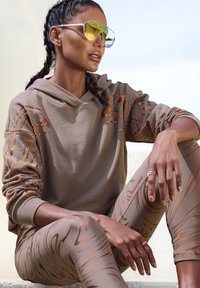 Brauner Hoodie mit orangefarbenen Mustern und passenden Leggings mit abstraktem Design. Das Model trägt reflektierende Sonnenbrillen und Ringe. Glatte Texturen.