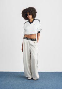 Λευκή αθλητική κοντή μπλούζα με μαύρες λεπτομέρειες και έμβλημα, συνδυασμένη με φαρδιά, κρεμ φόρμα με μαύρη μέση και ρίγες στα πλαϊνά.