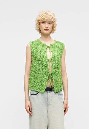 Ξανθό άτομο που φοράει αμάνικο πράσινο πλεκτό γιλέκο δεμένο με τρία φιόγκους, συνδυασμένο με ψηλόμεσο ανοιχτό μπλε τζιν και μαύρη ζώνη.