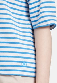 Swobodny sweter w poziome paski, z naprzemiennymi jasnoniebieskimi i białymi pasami, z zaokrąglonym dołem oraz małym logo w niebieskim haftem blisko dolnej krawędzi.