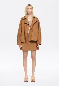 Bruine suède oversized jas met een uitgesneden kraag, gecombineerd met een bijpassende minirok. Het model draagt slip-on sandalen met een bloemenaccent.