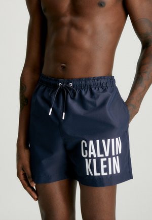 Bañadores de color marino hechos de un tejido suave, con una cinturilla elástica con cordón y un gran logo blanco de "CALVIN KLEIN".