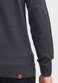 Pull gris foncé avec un ourlet et des poignets côtelés, des patchs aux coudes et une petite étiquette en cuir marron en bas. Texture lisse sur l'ensemble.