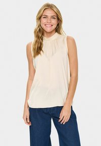 Donna che indossa un top senza maniche beige e jeans blu scuro, sorridente, con lunghi capelli biondi ondulati, in piedi davanti a uno sfondo bianco semplice.