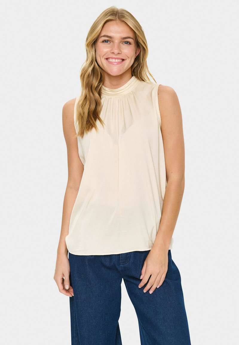 Donna che indossa un top senza maniche beige e jeans blu scuro, sorridente, con lunghi capelli biondi ondulati, in piedi davanti a uno sfondo bianco semplice.