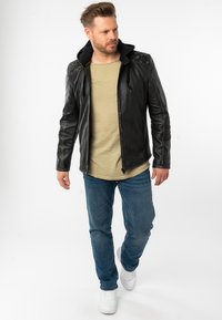 Schwarze Lederjacke mit Kapuze, strukturierten Schultern, einem Reißverschluss, kombiniert mit einem hellgrünen Hemd, blauen Jeans und weißen Turnschuhen.