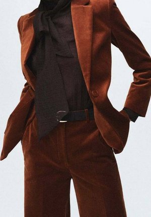 Kobieta w aksamitnym garniturze w kolorze rdzy, czarna bluzka z dużą kokardą przy szyi, z rękami w kieszeniach.