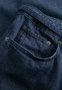 Κοντινή λήψη τσέπης από σκούρο μπλε denim τζιν με ορατές ραφές και μεταλλικά νύχια στις άκρες του υφάσματος.
