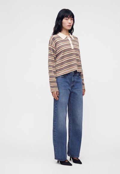 Femme debout portant une chemise rayée à manches longues avec col, un jean bleu à jambes larges et des talons noirs à bouts pointus sur un fond uni.