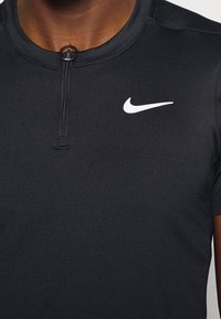 Svart atletisk skjorta med svart halvmatchning krage bärs av en person, med en vit Nike swoosh-logotyp på vänster bröst.