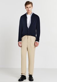 Marineblauer Cardigan, zugeknöpft, über einem weißen Hemd mit Kragen. Beigefarbene, plissierte Hosen und schwarze Schuhe. Glatter, strukturierter Stoff.