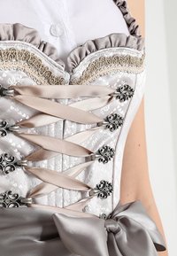 Corset gris clair à motifs floraux, laçage en ruban de satin, accents argentés et un nœud en soie. Présente des bords froncés au niveau de l'encolure.