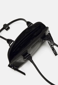 Sac en cuir noir à bandoulière avec une fermeture éclair ouverte et des poches intérieures visibles. Caractérisé par des sangles élégantes et un logo de marque en relief sur l'extérieur.