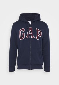 Tmavě modrá bavlněná mikina na zip. Má podšitou kapuci, dvě přední kapsy a výrazný nápis „GAP“ v červené a bílé barvě. Hladká textura.