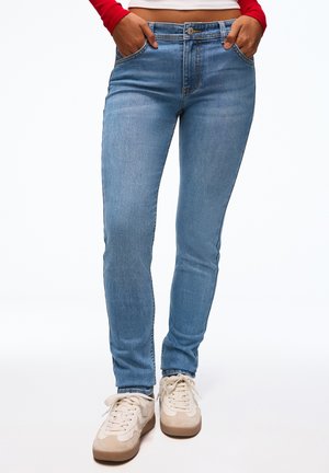 Personne portant un jean skinny bleu clair, des baskets beige et blanches, avec les mains dans les poches avant et un haut rouge à manches longues partiellement visible.