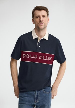 Homme aux cheveux courts portant un polo bleu marine avec un col blanc et une bande horizontale rouge indiquant "POLO CLUB", debout les mains dans les poches.