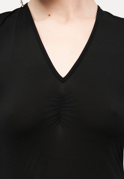 Μαύρο τοπ με V λαιμόκοψη και ρυτιδωτή λεπτομέρεια στη λαιμόκοψη, φορεμένο από πρόσωπο με ακάλυπτο λαιμό και ανώτερο στήθος.